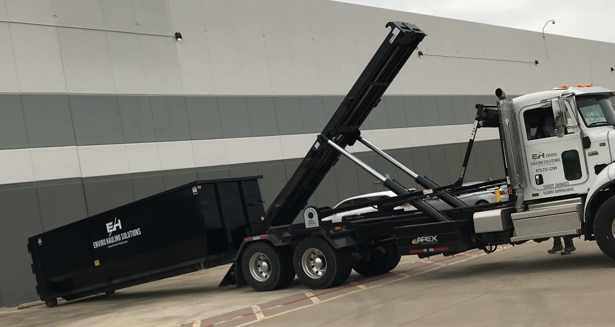 EnviroHauling roll-off truck loading a black open-top container beside a warehouse
